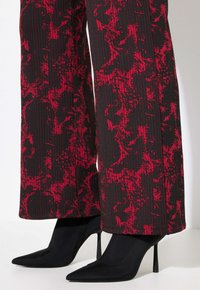 Schwarze High-Heels-Stiefel mit spitzer Zehe, aus einem glatten Material, kombiniert mit weit geschnittenen Hosen in Rot und Schwarz mit strukturiertem, marmoriertem Muster.