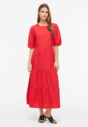 Nő áll, élénk vörös réteges midi ruhát visel, puffadt könyékig érő ujjal és fekete nyitott orrú magas sarkú papucsban.