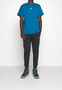 Modrá bavlněná tričko s bílým logem Nike, doplněné černými tepláky s barevnou nášivkou a vysokými zeleno-černými teniskami.