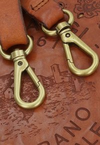 Detalle de bolsa de cuero marrón con herrajes de latón y una superficie texturizada, con patrones en relieve y elementos numéricos visibles.