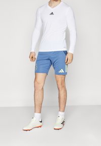 Valkoinen pitkähihainen urheilupaita mustalla logolla, siniset shortsit keltaisin yksityiskohdin ja logolla sekä valkoiset jalkapallokengät oranssein yksityiskohdin.