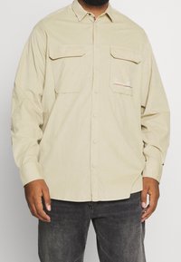 Beżowa bawełniana koszula z długim rękawem, z dwoma kieszeniami na piersi, zapięciem na guziki z przodu oraz haftowanym logo na lewej kieszeni. Tkanina o fakturze i luźnym kroju.