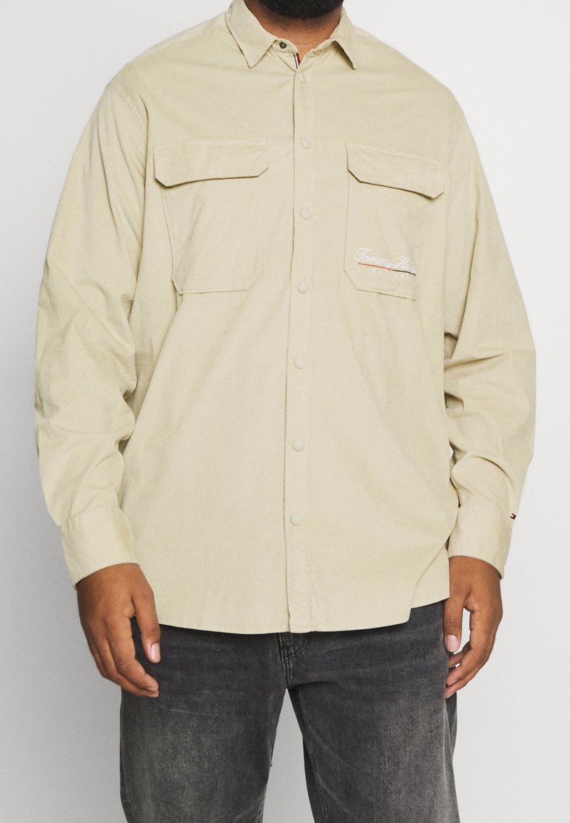 Beżowa bawełniana koszula z długim rękawem, z dwoma kieszeniami na piersi, zapięciem na guziki z przodu oraz haftowanym logo na lewej kieszeni. Tkanina o fakturze i luźnym kroju.