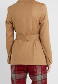 Jasnobrązowa marynarka z paskiem, o wyraźnych ramionach i gładkiej fakturze, z klasycznym kołnierzem i czystymi liniami, połączona z spodniami w kratę.