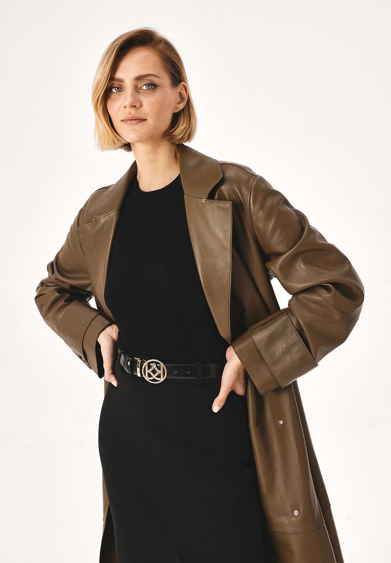 Oliven grøn læder trench coat med en bred krave, parret med en sort tætsiddende kjole og bælte med en rund metalspænde.