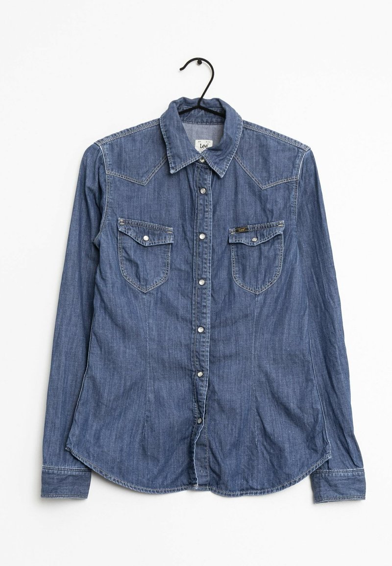 Chemise en denim bleu à manches longues avec boutons et deux poches poitrine sur un cintre noir, fond blanc.