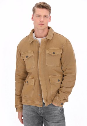 Ung mand iført en beige knappet jakke over en hvid skjorte, med den ene hånd i lommen, stående mod en hvid baggrund.