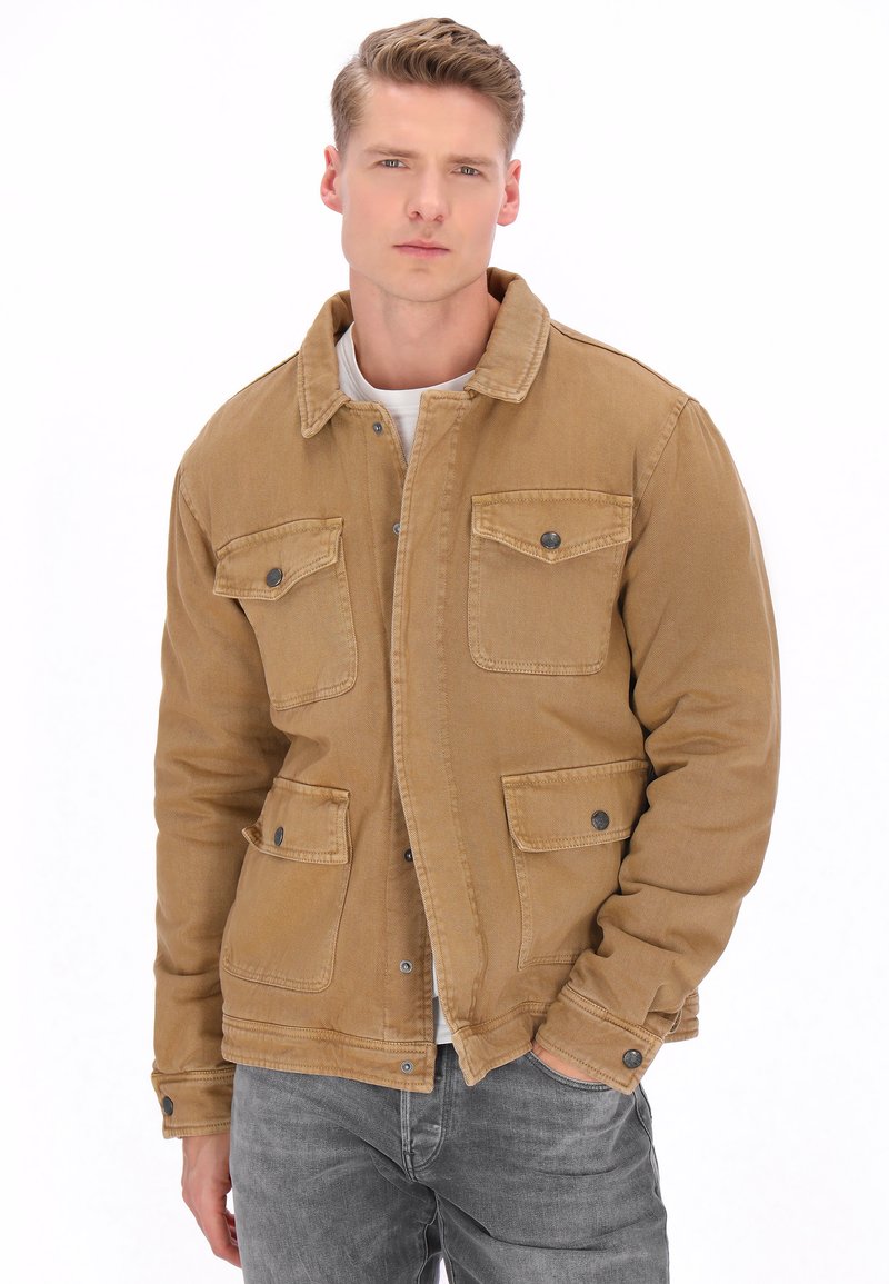 Jeune homme portant une veste beige boutonnée par-dessus une chemise blanche, avec une main dans la poche, debout devant un fond blanc.