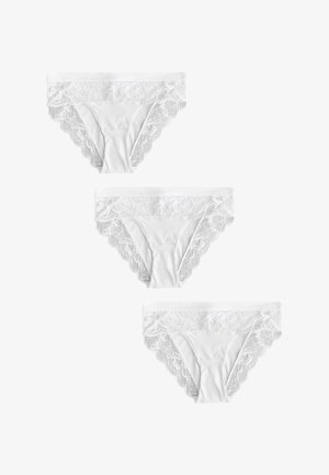 Frau trägt weißen BH mit Spitzenbesatz und passende Spitzenverzierte Slips vor schlichtem Hintergrund.