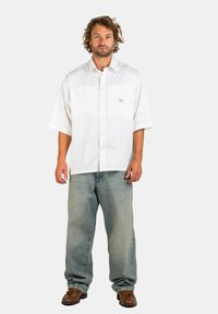 Mann mit lockigem Haar, der ein übergroßes weißes Hemd, lockere hellblaue Jeans und braune Lederschuhe trägt, vor einem schlichten Hintergrund stehend.