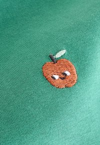 Grön tyg med en broderad brun äpple som har ett leende ansikte och ett litet grönt blad, vilket ger en lekfull touch till plagget.