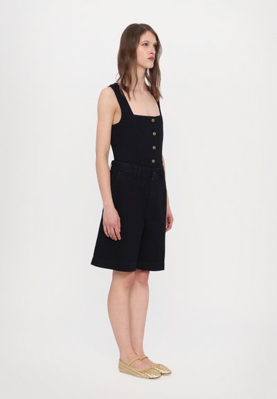 Black sleeveless jumpsuit with a square neckline, golden buttons, and matching shorts. Model wears gold woven flats. Simple, minimalistic design.