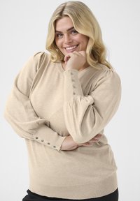Pull en maille beige avec des manches bouffantes, présentant des accents de boutons aux poignets et un col rond. Texture douce, coupe décontractée.