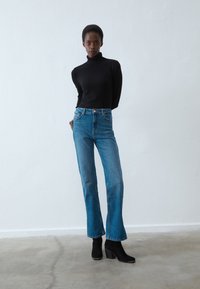 Svart polo genser, blå høytlivede flagrende jeans og svarte helte støvler. Glatt tekstur og tettsittende design med minimalt med detaljer.