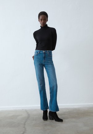 Svart polo genser, blå høytlivede flagrende jeans og svarte helte støvler. Glatt tekstur og tettsittende design med minimalt med detaljer.