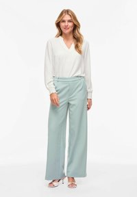 Femme debout, portant un chemisier blanc à manches longues, un pantalon large bleu clair et des sandales à talons roses claires.