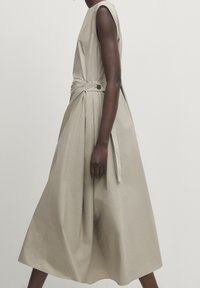 Personne à la peau foncée portant une robe midi beige sans manches avec des boutons sur le côté et des plis, se tenant devant un fond clair uni.