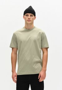 Tricou din bumbac de culoare verde măsliniu, cu m�âneci scurte și gât rotund, având un mic logo alb. Purtați cu pantaloni negri și o căciulă neagră.