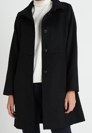 Manteau noir mi-long avec des boutons, porté sur un pull à col roulé blanc et un pantalon sombre, présenté sur une personne debout.