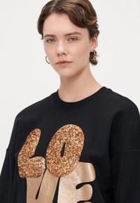 Μαύρη μπλούζα με μεγάλα χρυσά και γυαλιστερά γράμματα "ΑΓΑΠΗ", με στρογγυλή λαιμόκοψη και άνετη εφαρμογή. Ο απαλός υφασμάτινος χαρακτήρας είναι ορατός.