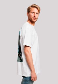 Blond man wearing a white T-shirt with a colorful mountain and forest graphic on the back, standing against a plain light background.