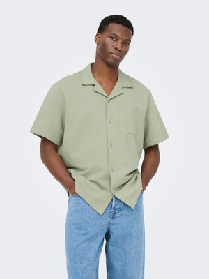 Homme portant une chemise à manches courtes vert clair avec une poche poitrine et un jean bleu clair, debout les mains dans les poches.