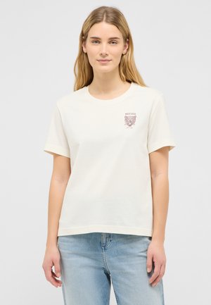 Young woman with long blonde hair wearing a white t-shirt with a small brown leopard graphic and light blue jeans, standing against a plain background.