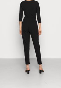 Svart jumpsuit med trekvartslånga ärmar, avsmalnande ben och dragkedja i ryggen. Slät tyg med en figurnära design och inga synliga mönster.