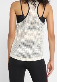 Mujer vistiendo una camiseta sin mangas de malla blanca sobre un sujetador deportivo negro y leggins negros, vista desde atrás.