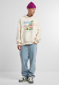 Krémová mikina s barevným grafickým potiskem, který zobrazuje palmy a budovy, v kombinaci se světle modrými džíny a zeleno-bílými teniskami.