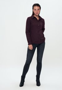 Camicia bordeaux a maniche lunghe con bottoni frontali, colletto classico, e jeans skinny scuri abbinati a stivali neri alla caviglia.