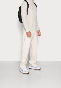 Ljust grå kabelstickad tröja, krämfärgade vida byxor och vita sneakers med svarta och bruna sulor. Svart ryggsäck bärs på ena axeln.