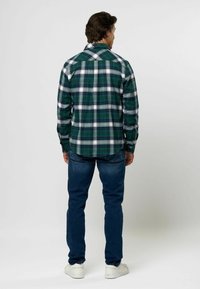 Man som står med ryggen vänd mot kameran, iklädd grön- och vitrutig skjorta, blå jeans och vita sneakers mot en enkel ljus bakgrund.