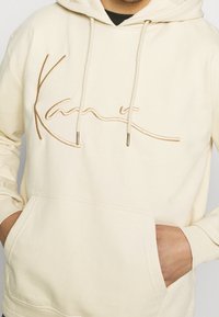 Krémová mikina s vyšitým béžovým podpisovým logem. Obsahuje přední kapsu a stáhnutí s kovovými špičkami. Měkká tkanina a dlouhé rukávy.