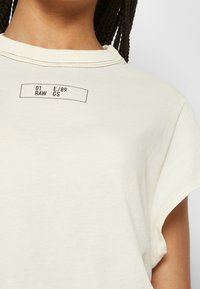 Krémové tričko s krátkým rukávem vyrobené z měkké tkaniny. Na přední straně se nachází malá tmavě potištěná nálepka s textem "01 RAW E/89 GS", jednoduchý design.