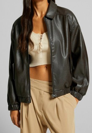 Kvinde iført en mørkebrun læderjakke over en beige ribbet crop top og beige plisserede bukser, stående med den ene hånd i lommen.