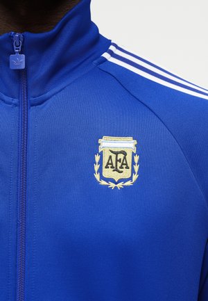 Mėlynas sportinis švarkas su baltomis trimis juostelėmis ant peties, aukštu apykakle užtrauktuku ir siuvinėtu AFA herbu ant krūtinės.