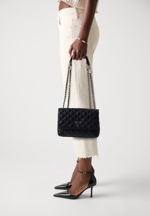 Sac à main matelassé noir avec un logo triangulaire, doté d'une bandoulière en chaîne et d'une fermeture à rabat. Porté avec un jean couleur crème et des talons aiguilles noirs.