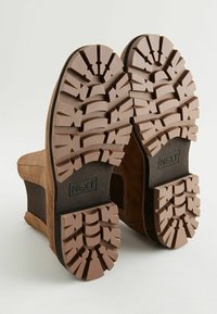 Botas de tobillo de ante marrón con suelas de goma que presentan profundos surcos. Los paneles laterales están hechos de un tejido texturizado. Presencia de la marca en la suela.