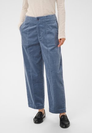 Pantalon en velours côtelé bleu taille mi-haute à jambes larges avec poches avant, porté avec des mocassins noirs et un haut beige à manches longues, mains du mannequin dans les poches.