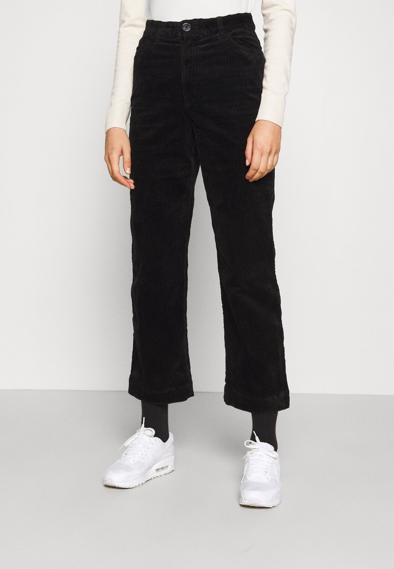 Personne portant un pantalon en velours côtelé noir, des chaussettes noires, des baskets blanches et un haut à manches longues de couleur claire, debout sur un sol gris.