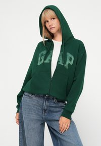 Zelena dukserica na zip s prednjim džepom i velikim "GAAP" logotipom. Dizajn s kapuljačom, rebrasti rukavi, izrađena od mekanog materijala. Nosi se preko bijele majice.