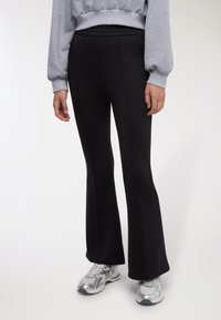 Pantaloni neri a zampa con tessuto liscio ed elastico. Presentano una vita alta e cuciture verticali, abbinati a scarpe da ginnastica grigie.