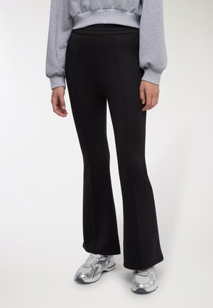 Pantaloni neri a zampa con tessuto liscio ed elastico. Presentano una vita alta e cuciture verticali, abbinati a scarpe da ginnastica grigie.