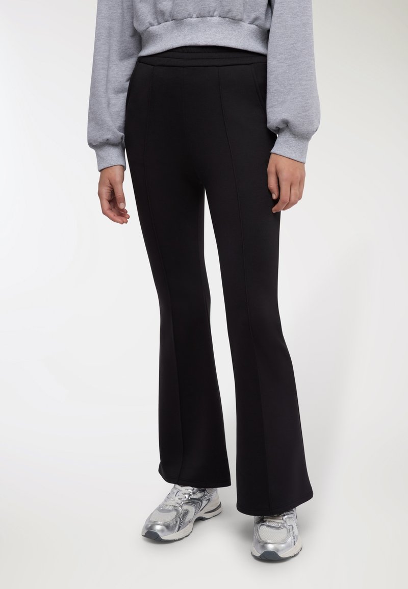 Pantaloni neri a zampa con tessuto liscio ed elastico. Presentano una vita alta e cuciture verticali, abbinati a scarpe da ginnastica grigie.