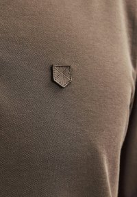 Hnedá textúrovaná látková košeľa s malým, tmavým vyšitým logom v tvare štítu na ľavej strane. Dizajn je jednoduchý a nenápadný.