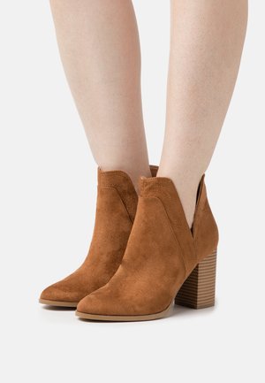 Brown suede ankle boots with a pointed toe, cut-out sides, and a stacked heel featuring horizontal grooves. Textured surface and matte finish.