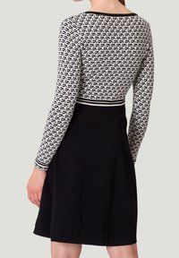 Langärmliges Kleid mit einem schwarz-weißen gemusterten Oberteil mit geometrischen Designs und einem einfarbig schwarzen Rock. Glatter Stoff und taillierte Passform.