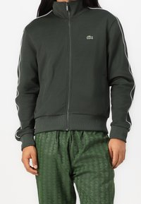 Chaqueta de cremallera verde oscuro con cuello alto, con franjas blancas a los lados y un logo de Lacoste. Material suave y puños acanalados. Combinada con pantalones verdes estampados.