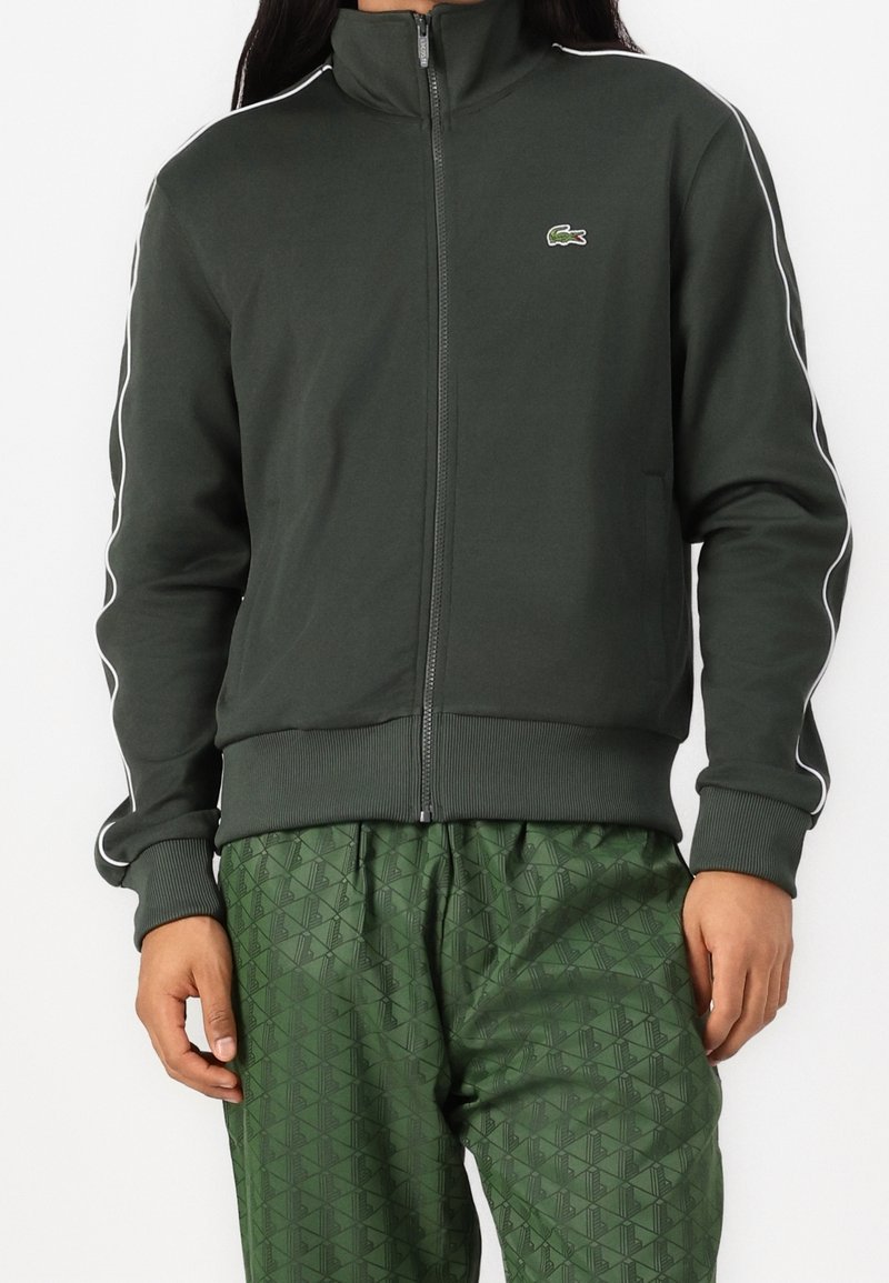 Chaqueta de cremallera verde oscuro con cuello alto, con franjas blancas a los lados y un logo de Lacoste. Material suave y puños acanalados. Combinada con pantalones verdes estampados.